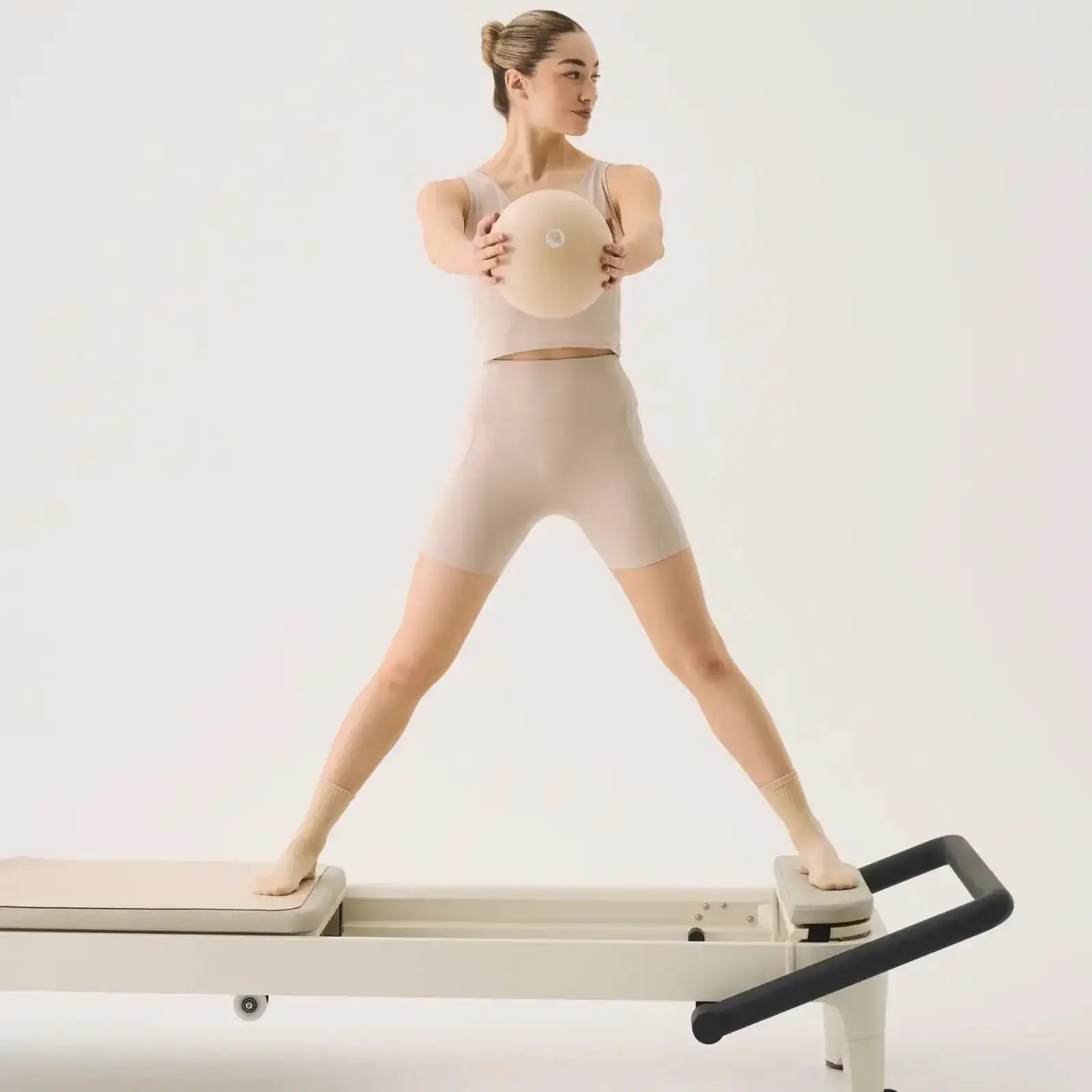 Female in almond gym gear balancing on a Reformer bed holding in her hands Almond Pilates ball