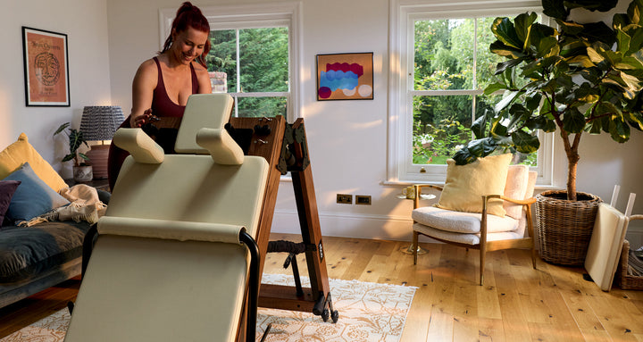 Woman adjusting a fitness reformer in a bright, cozy living room with plants and stylish decor.