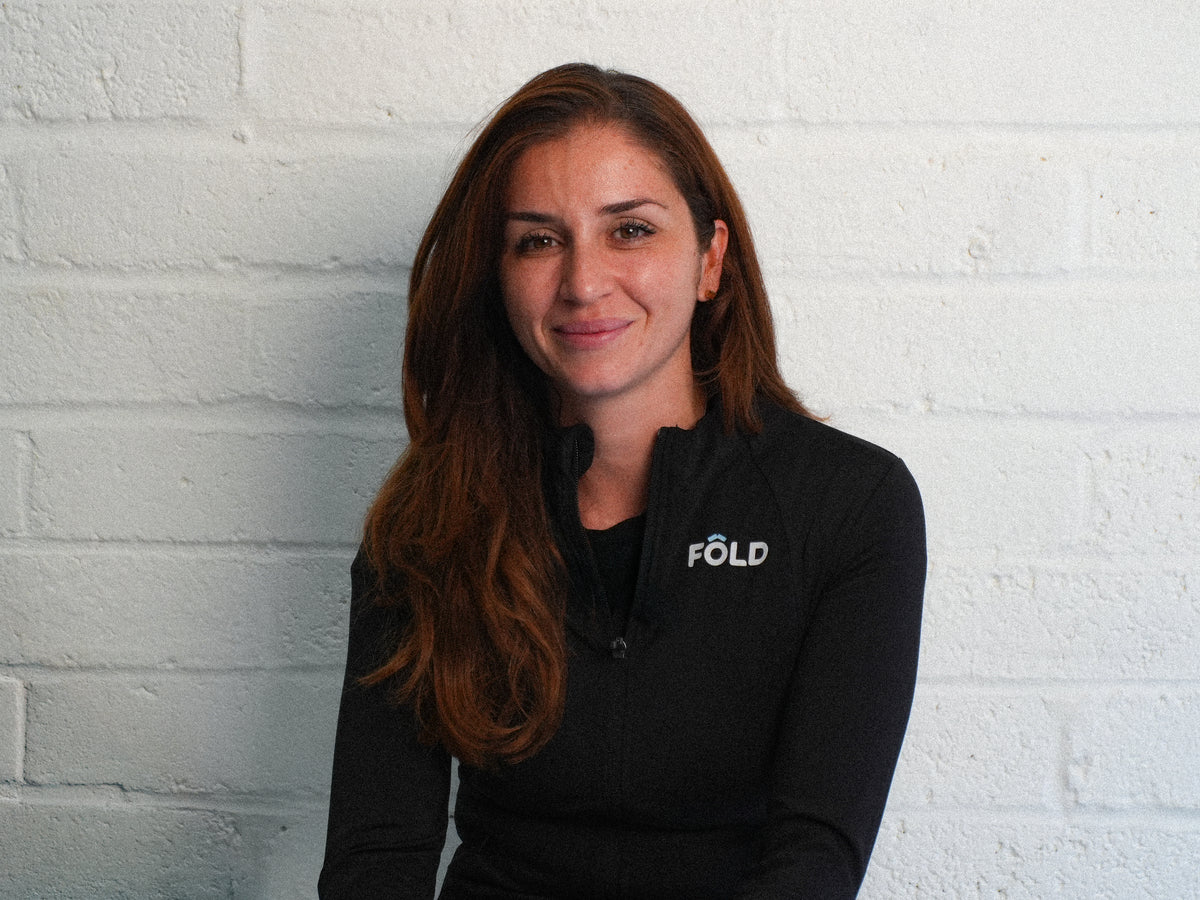 Portrait of a woman in a black FÖLD jacket, smiling in front of a white brick wall.