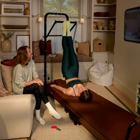 Woman exercising on a tower reformer machine in a living room with another person observing.