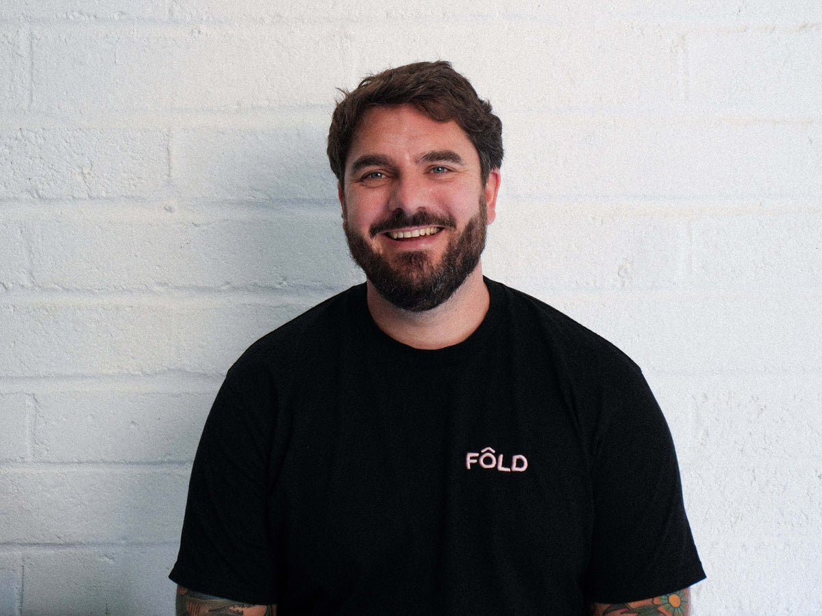 Smiling man with a beard wearing a black T-shirt with 'FÔLD' logo against a white brick wall.