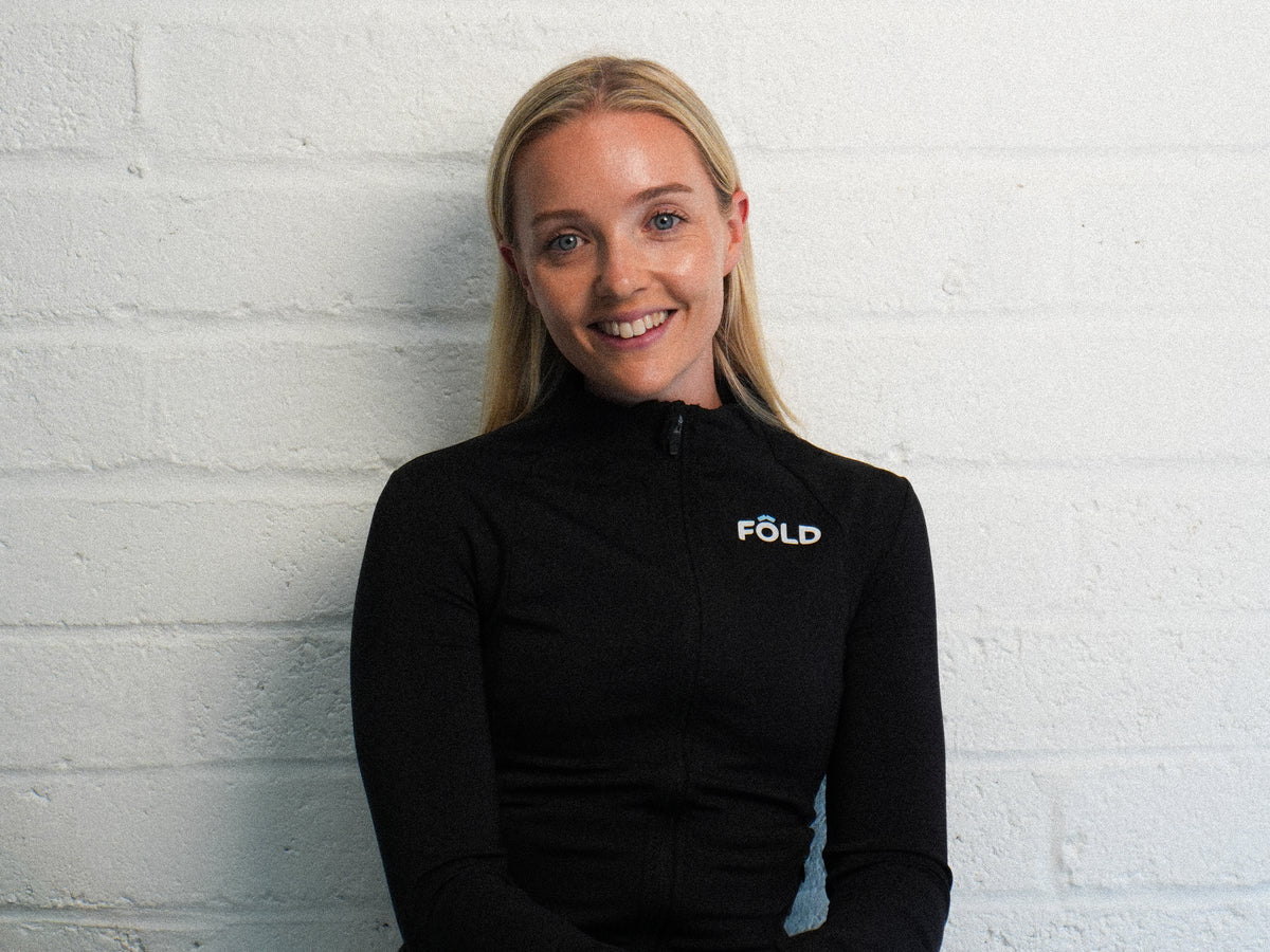 Smiling woman in a black zip-up jacket with FOLD logo, seated against a white brick wall.