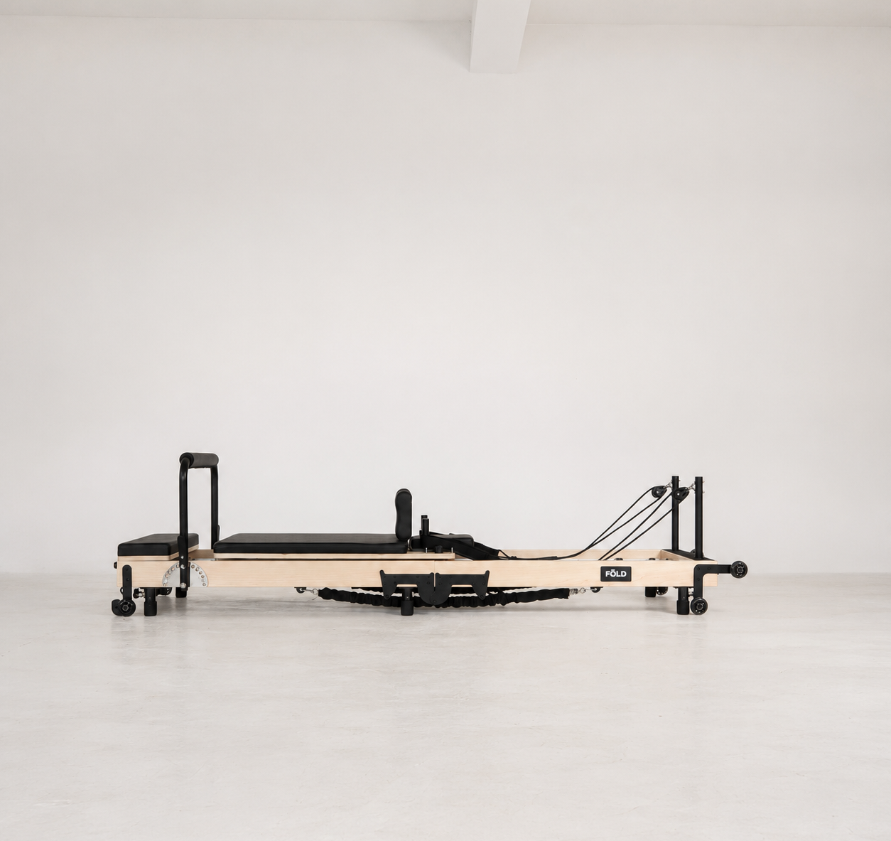 A Pilates reformer machine with a light natural wood frame and black padded components, shown in profile against a clean white studio background.
