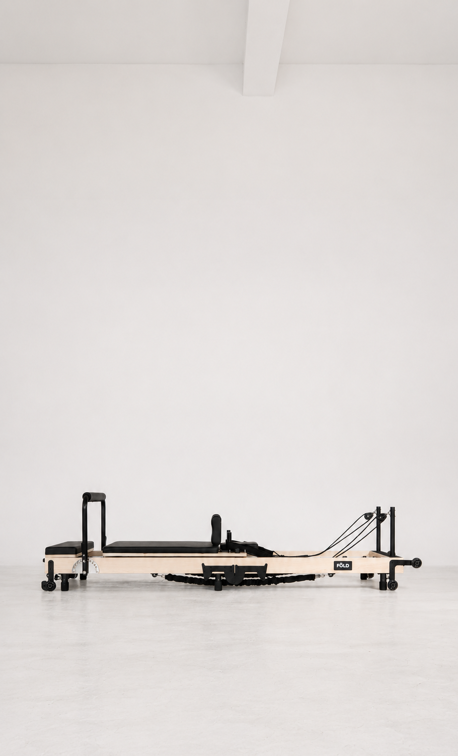 A Pilates reformer machine with a light wood frame and black padding, positioned horizontally against a minimalist white studio background.