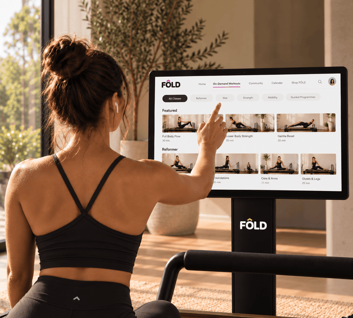 Woman exercising indoors, interacting with FOLD fitness screen showcasing on-demand workouts and classes.