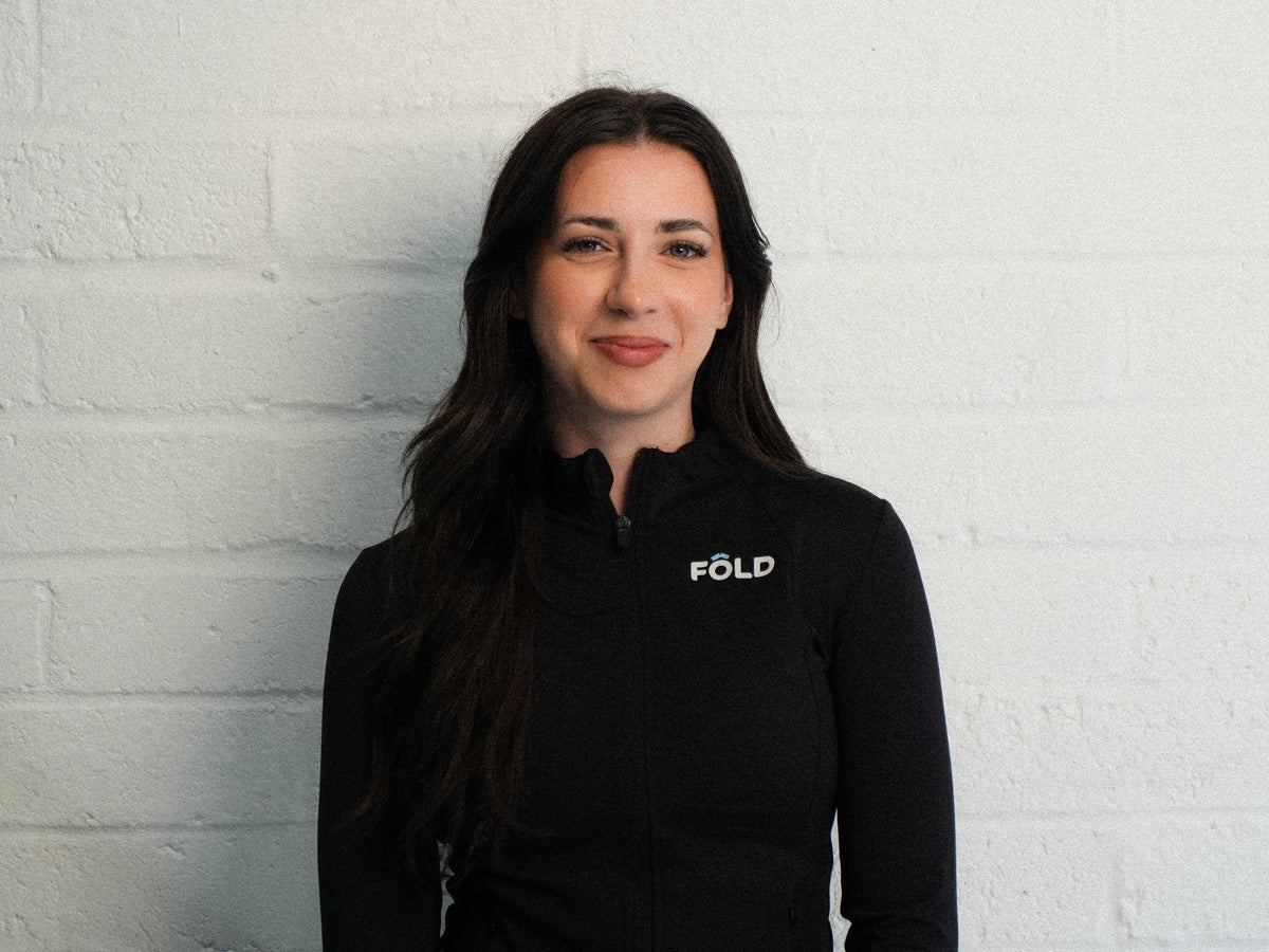 Smiling woman in black FOLD jacket against a white brick wall, showcasing a sporty and professional look.