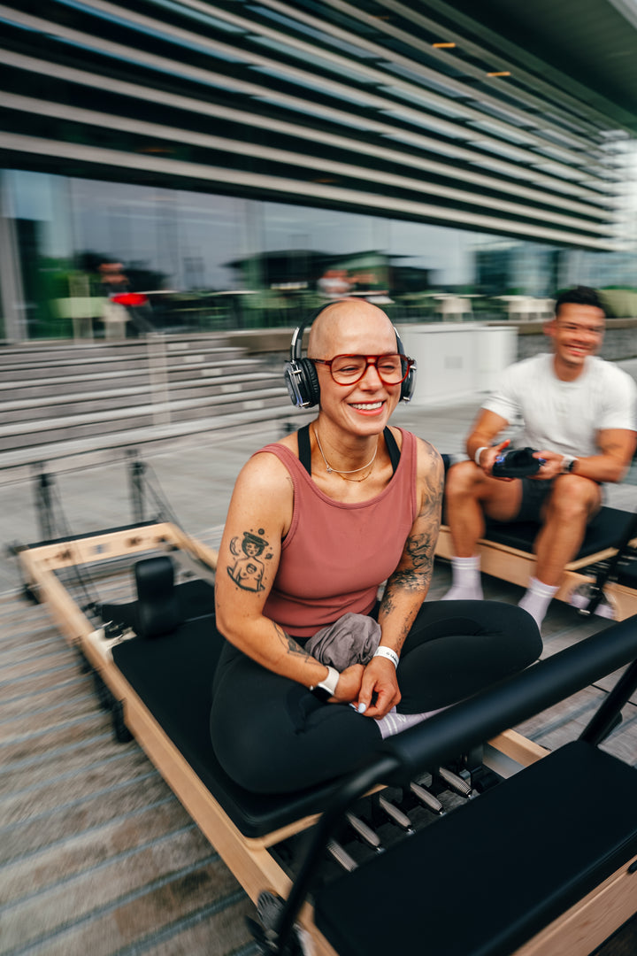 Woman with tattoos and headphones smiles during a fitness class on a reformer, showcasing joy and enthusiasm.